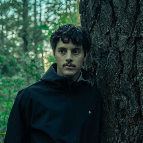 Daryn Steytler, founder of LINKED, leans against a tree trunk in a dark jacket amid forest greenery.